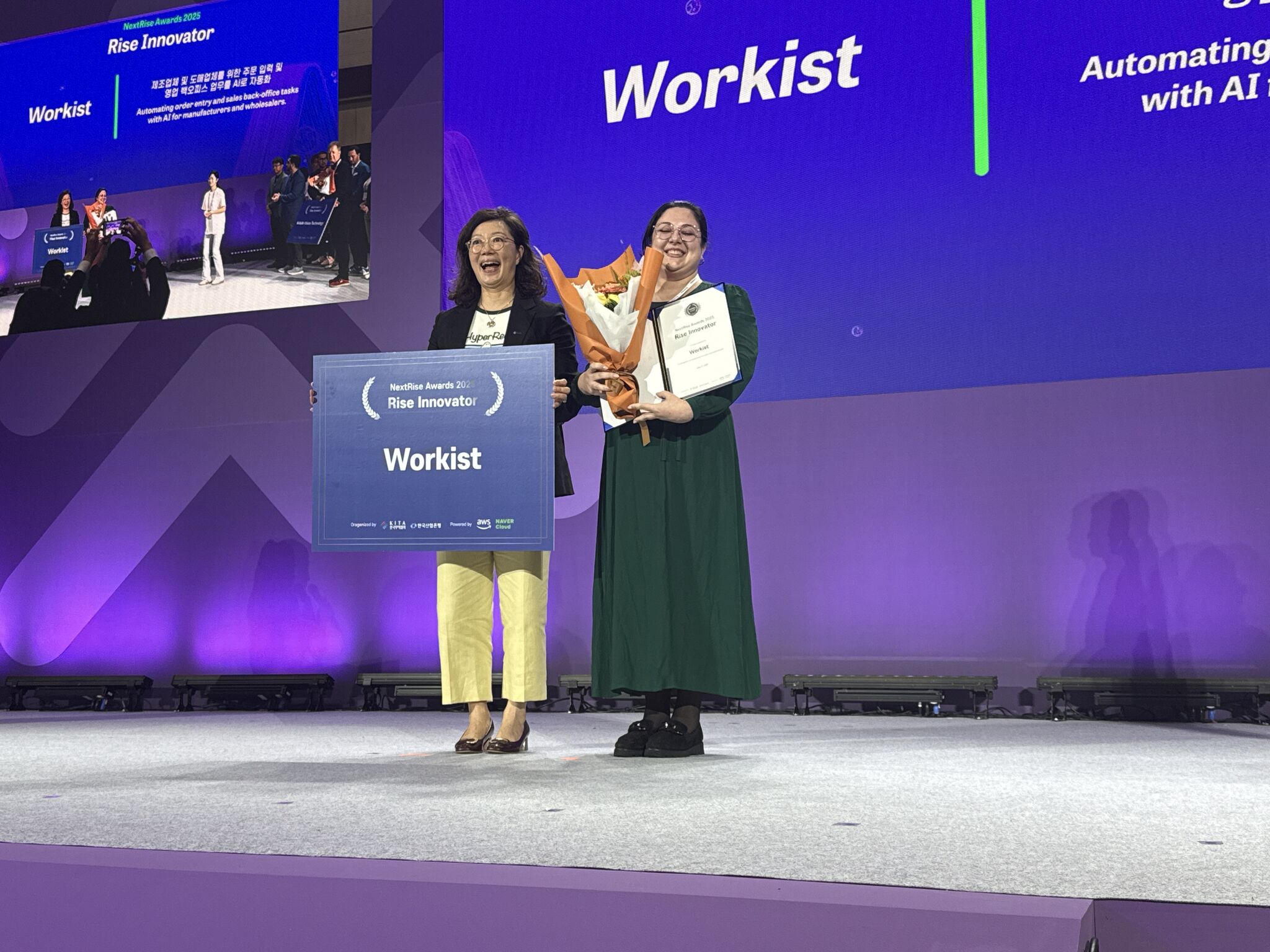 Workist receiving Rise Innovator Award – A woman holds flowers, a certificate, and a large award sign reading “Workist” while smiling on stage with another presenter at NextRise 2025.